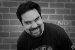 A man with dark hair and a goatee smiles while looking to the side against a brick wall.