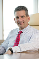 A man with short dark hair wearing a light striped shirt and a red patterned tie, sitting at a desk.