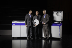 Three men in suits stand between two pieces of industrial equipment, one holding a reel.