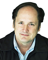A man with dark hair and a light blue shirt wearing a black jacket against a white background.
