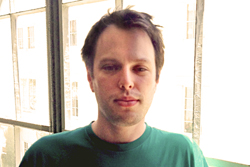 A man with short brown hair wearing a green t-shirt stands before a large window.