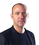 A bald man with fair skin and a serious expression wears a dark jacket over a black shirt.