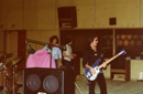 A musician plays a blue electric bass guitar on a stage with other band members in the background.