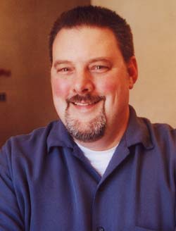 A smiling man with short dark hair and a salt-and-pepper goatee wearing a blue button-down shirt.