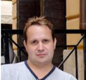 A man with short brown hair wearing a light grey v-neck shirt.