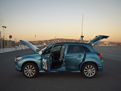 A blue hatchback parked on an asphalt lot with its hood, doors, and trunk open against a sunset sky.