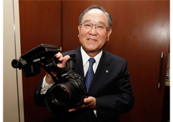 An elderly man in a dark suit and tie holds a professional DSLR camera and a video camcorder.