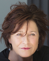 A close-up portrait of a woman with wavy, reddish-brown hair and light-colored eyes looking toward the camera.
