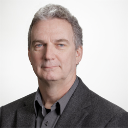 A middle-aged man with graying hair and blue eyes wearing a gray shirt and dark blazer.