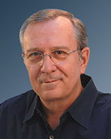 A man with short light hair and glasses wearing a dark blue collared shirt against a blue background.