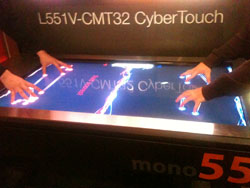 Four hands are shown interacting with a large, illuminated touch screen display that shows abstract graphics and text.