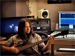 A man with long hair plays a bass guitar in a music studio with monitors and recording equipment.