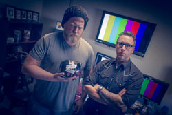 Two men stand in a studio; one holds a helmet while the other stands with arms crossed near color bar monitors.