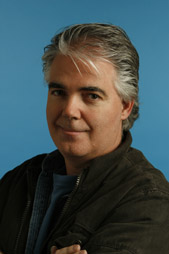 A man with salt-and-pepper hair and a dark jacket smiling against a solid blue background.