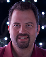 A smiling man with dark hair and a goatee wearing a maroon shirt against a dark, bokeh background.