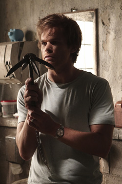 A man in a light t-shirt holds a black pickaxe with both hands in a rustic, dimly lit room.