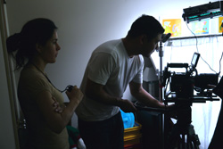 A man in a white t-shirt adjusts a professional camera on a tripod while a woman watches nearby.