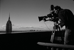 A person operates a large professional video camera on a rooftop overlooking a city skyline.