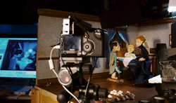 A digital camera on a tripod filming small puppets in a miniature set.