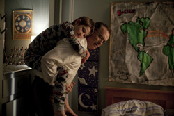 A man bends forward while a young child clings to his back in a dimly lit room.