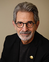 A man with gray hair, a salt-and-pepper beard, and round glasses wearing a black blazer.