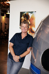 A smiling woman in a dark blue shirt and jeans leans against a silver metallic object near a poster.