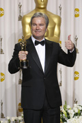 A man in a black tuxedo holding an Oscar statuette and giving a thumbs up in front of a gold Oscar backdrop.