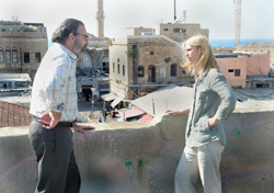 A man in a white shirt and a woman in a grey shirt talk on a rooftop overlooking a cityscape with minarets.