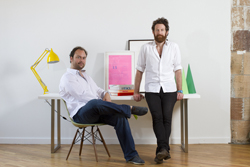 Two men in white shirts pose in a studio with a desk, yellow lamp, and colorful artwork.