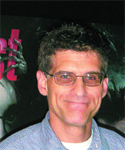 A man with short dark hair and glasses smiling in front of a dark poster.
