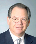 A man with light skin and brown hair wearing glasses, a white shirt, and a dark suit jacket.