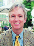 A man with light hair smiling, wearing a patterned blazer, blue shirt, and yellow tie.