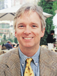 A smiling man with light hair wearing a tan blazer, blue shirt, and yellow patterned tie.