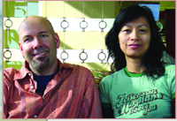 A man in an orange shirt and a woman in a green t-shirt sitting together indoors.