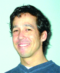 A smiling man with dark, wavy hair wearing a dark blue sweater against a light background.