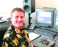 A smiling man in a black floral shirt sits at a desk with two monitors, a keyboard, and studio speakers.