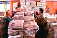 Three people stand and talk in a room filled with large stacks of newspapers.