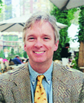 A smiling man with blond hair wearing a patterned blazer, blue shirt, and yellow patterned tie.