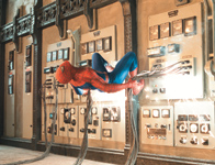 A person in a red and blue superhero suit clings to a large electrical control panel with sparks flying.
