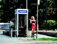 A blonde woman in a black top and red skirt stands beside a silver telephone booth outdoors.