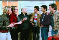 Six men standing in a row, talking and holding glasses of red wine.