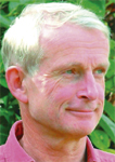 A fair-skinned man with short light hair looking slightly to the side against a green leafy background.