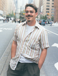 A man wearing a tan and white striped button-down shirt over a white t-shirt stands on a city street.
