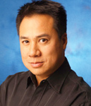 A man with short dark hair wearing a black button-down shirt against a blue background.