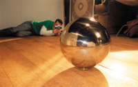 A person lying on a wooden floor, aiming a camera at a large, shiny metallic sphere.