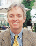 A smiling man with light hair wearing a tan blazer, blue shirt, and yellow patterned tie.