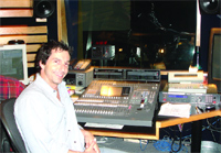 A man smiles while sitting in front of a large audio mixing desk and studio equipment.