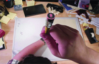 A person's hand holds a yellow marker over a large open notebook on a cluttered desk.