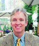 A smiling man with blond hair wearing a patterned blazer, blue shirt, and gold tie.