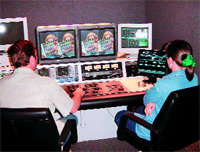 Two people sit at a desk facing multiple computer monitors displaying video and technical data.